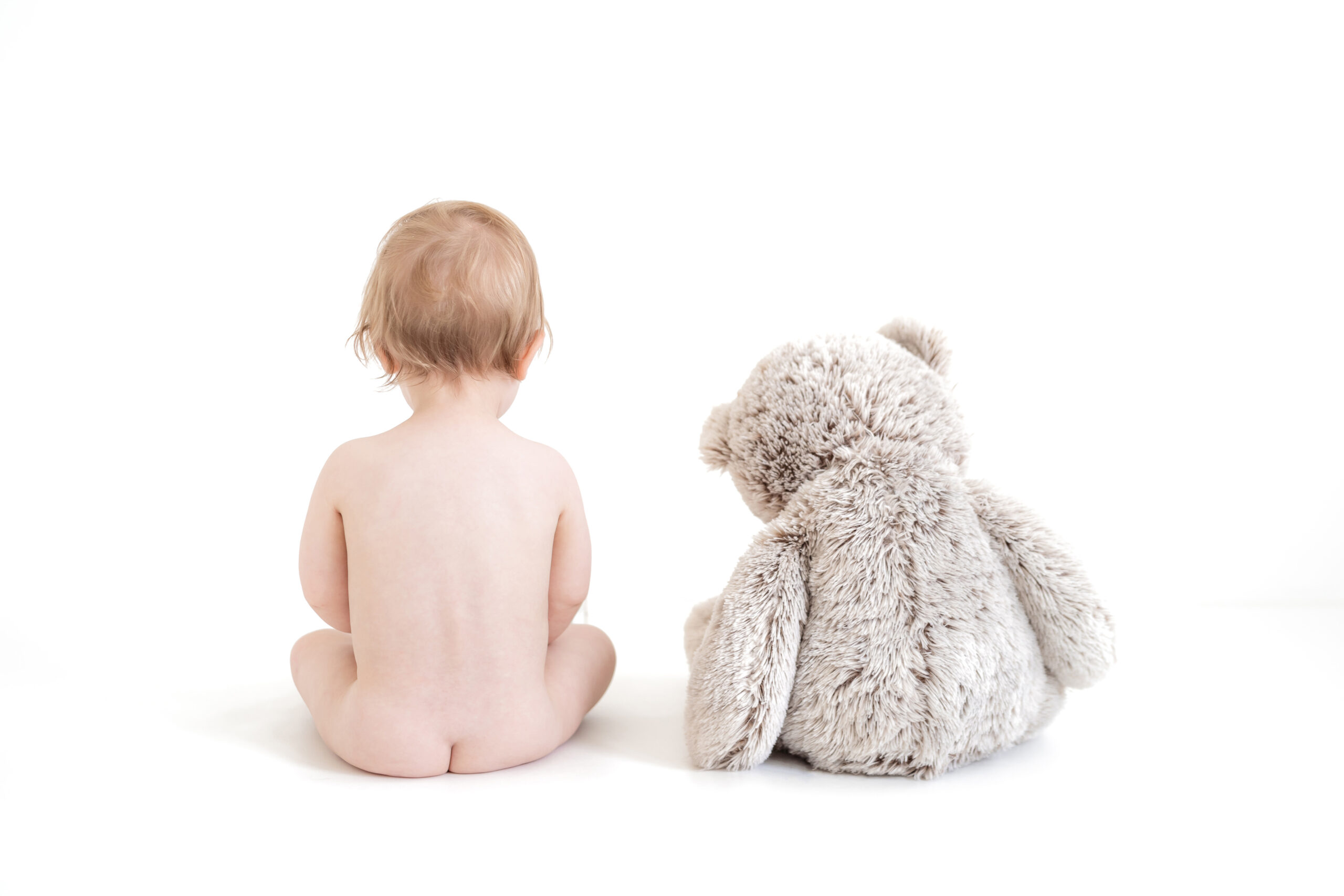 Baby and Teddy Bear sat with their backs towards the camera against a white background.