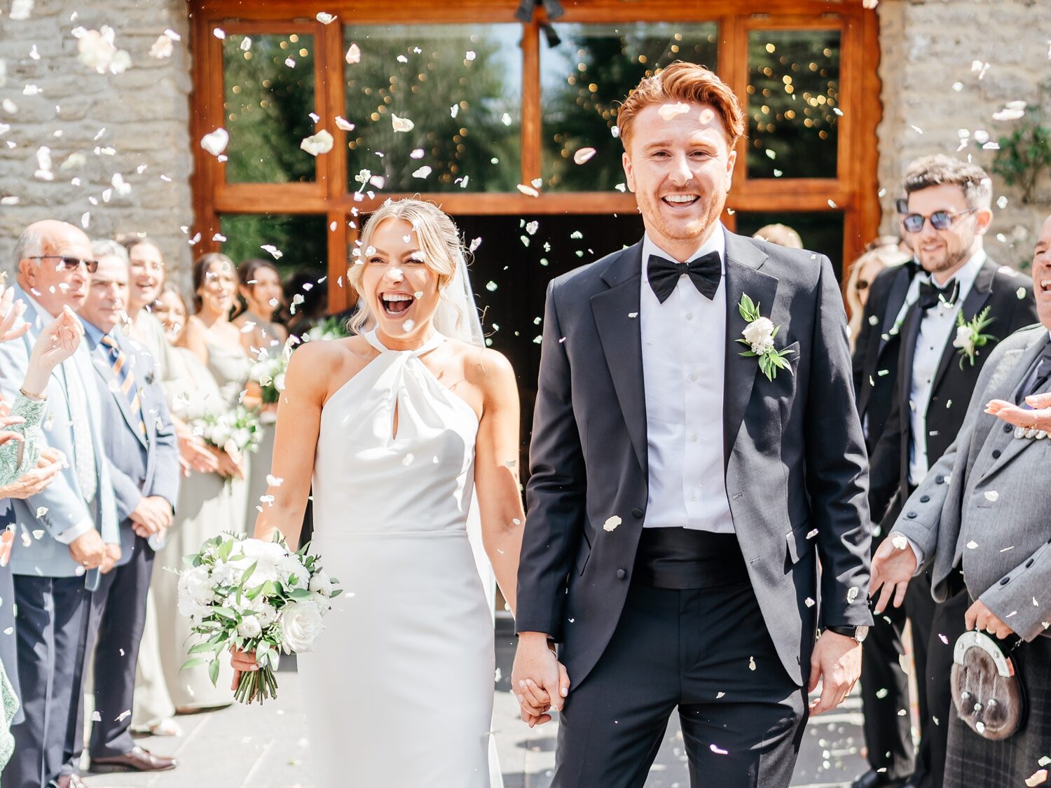 A joyful bride and groom walk hand-in-hand, smiling amid a shower of confetti. They are surrounded by cheering guests, radiating happiness at Wick Farm Bath in Somerset