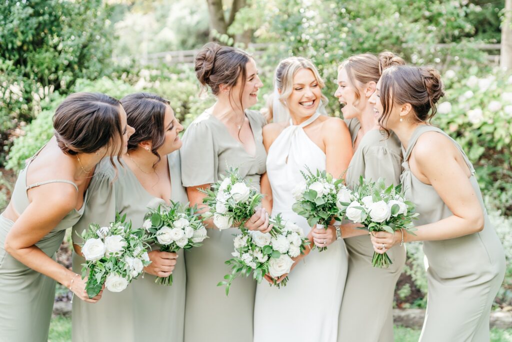 A joyful bride in a white gown (Portia by Love Spell Design) stands with five bridesmaids in sage green bridesmaid dresses. They hold white rose bouquets, laughing together in a lush garden setting at Wick Farm Bath Wedding Venue.