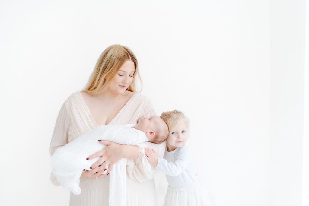 Mum holding her baby by the studio window during a relaxed baby photography session with big sister in Westbury.